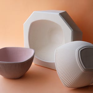 May include: A studio shot of three ceramic objects. A mauve bowl with vertical ridges, a white geometric form, and a white, rounded object with a ridged surface. The objects are set against a peach-colored background.