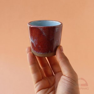 May include: A small, handmade ceramic cup with a deep red exterior and a light blue interior. The cup has a rustic, textured finish and a natural, unglazed base. The cup is held in a hand against a peach-colored background.