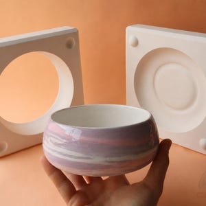 以下が含まれることがあります： ピンク、白、ラベンダーの渦巻き模様のセラミックボウル。円形の切り抜きとくぼみのある2つの白い正方形の型が背景にあります。ボウルは手で持たれています。