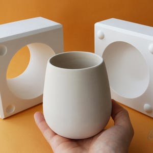 Peut inclure: Deux moules en plâtre blanc pour la fabrication d'une tasse en céramique. Les moules sont carrés et ont une ouverture ronde au centre. Une tasse en céramique blanche est posée sur une main, devant les moules.