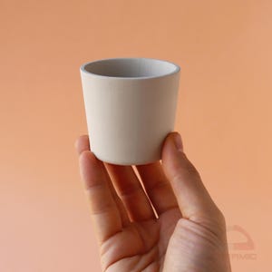 Könnte beinhalten: Eine kleine, cremefarbene Keramiktasse mit einem hellgrauen Rand. Die Tasse hat eine zylindrische Form mit einem leicht konischen Körper. Der Hintergrund ist einfarbig pfirsichfarben. Die Tasse wird in einer Hand gehalten.