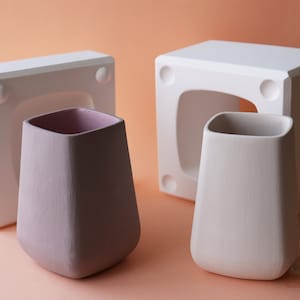 Peut inclure: Deux vases en céramique, l'un blanc et l'autre violet clair, sont présentés à côté de leurs moules. Les moules sont en plâtre blanc et ont une forme carrée avec une ouverture arrondie.