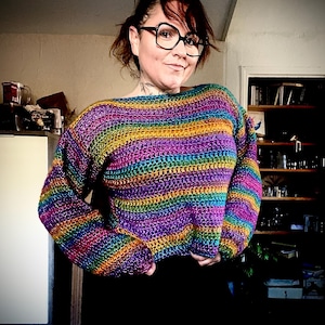 Peut inclure: Un pull crocheté à rayures arc-en-ciel avec des manches longues. Le pull présente des bandes horizontales de différentes couleurs, notamment le violet, le bleu, le vert, le jaune et le rouge. Le pull est de style court.