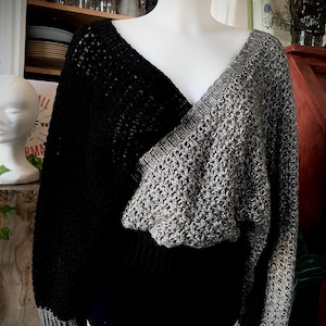 Peut inclure: Un pull en crochet bicolore noir et gris, présenté sur un mannequin. Le pull présente une encolure en V avec des manches longues et un motif de blocs de couleurs contrastés. Les manches et l'ourlet ont une bordure grise.