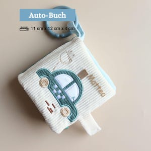 Könnte beinhalten: Ein weißes Stoffbuch aus Cord mit einem gestickten grünen Auto, dem Namen Matteo und einem blauen Kunststoffring. Das Buch misst 11 cm x 12 cm x 4 cm. Der Text Auto-Buch ist oben im Bild zu sehen.