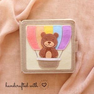 Può includere: Un libro per bambini in feltro beige fatto a mano con un design a mongolfiera colorato. Il pallone presenta un arcobaleno di colori e un orsacchiotto marrone. Il libro ha un bordo beige e una piccola chiusura a linguetta. Il testo "handcrafted with" e un cuore sono in basso.