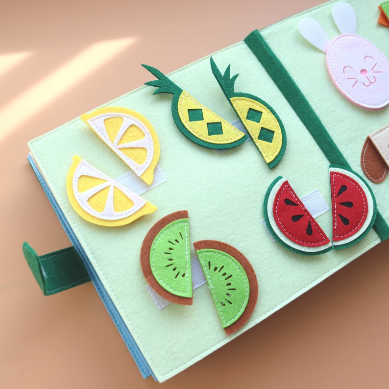 May include: A green felt book featuring various fruit shapes, including lemons, pineapples, kiwi, and watermelon. The fruit shapes are attached with velcro and can be moved around.