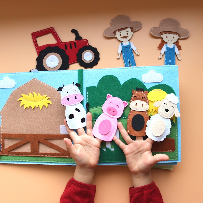 May include: A felt book featuring a red tractor, a barn, a cow, a pig, a horse, and a sheep. The book is open and a child's hands are holding the felt animals.