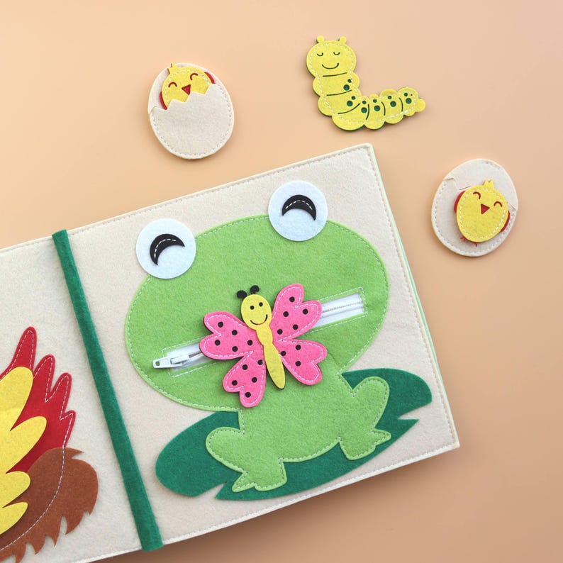 May include: A felt book for children featuring a green frog, a pink butterfly, a yellow chick, and a green caterpillar. The book has a zipper on the frog's mouth.