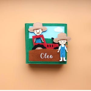 May include: A felt book featuring a red tractor, a farmer and a girl in overalls. The book has a green cover and the name Cleo is written on the front.