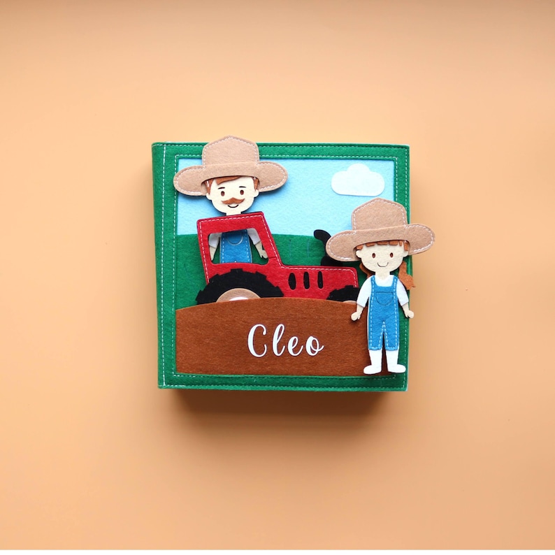 May include: A felt book featuring a red tractor, a farmer and a girl in overalls. The book has a green cover and the name Cleo is written on the front.
