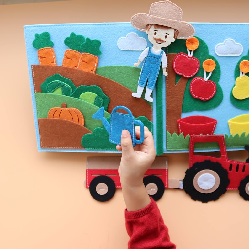 May include: A felt book for children featuring a farmer, a tractor, a watering can, carrots, pumpkins, apples, and pears. The book is open to reveal the different scenes.