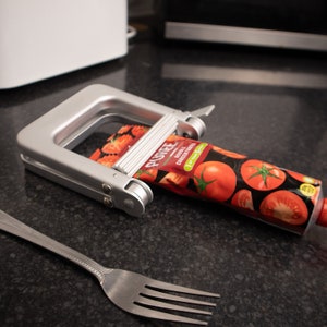 May include: A silver metal tube squeezer with a red and white tomato sauce tube. The tube has the text "PUGNEE DOUBLE CONCENTRATED" on it. The tube squeezer is on a black countertop with a silver fork.