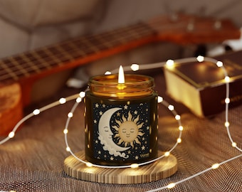Puede incluir: Una vela encendida en un tarro de vidrio con un diseño celestial que presenta una luna creciente y un sol sobre un fondo negro. La vela está sobre un posavasos de madera, rodeada de luces de cadena. El tarro tiene una tapa dorada.