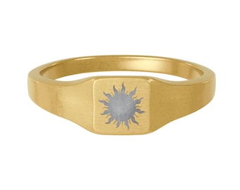 Puede incluir: Un anillo de sello dorado con una cara cuadrada que presenta un emblema de sol de plata cepillada grabado en el centro. La banda es lisa y pulida, mostrando un tono de oro amarillo consistente en toda la estructura circular de la pieza.