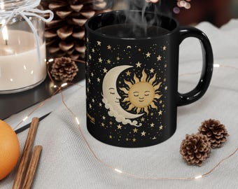 Puede incluir: Taza de cerámica negra con un diseño celestial que presenta un sol y una luna sonrientes, rodeados de estrellas doradas. La taza tiene un asa negra y contiene una bebida caliente. La taza está sobre una superficie gris claro.