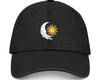 Puede incluir: Una gorra de béisbol negra con un diseño bordado en el panel frontal. El bordado representa una luna creciente blanca con nubes estilizadas en la parte inferior y un sol amarillo dorado con rayos radiantes situado junto a la luna.