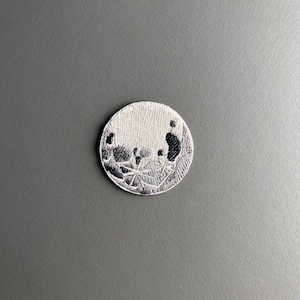 Könnte beinhalten: Ein runder, gestickter Aufnäher mit einem detaillierten Design in Weiß- und Schwarztönen. Der Aufnäher hat ein strukturiertes Aussehen, wobei aufwendige Stiche ein einzigartiges Muster erzeugen. Der Aufnäher ist wahrscheinlich für Kleidung oder Accessoires.