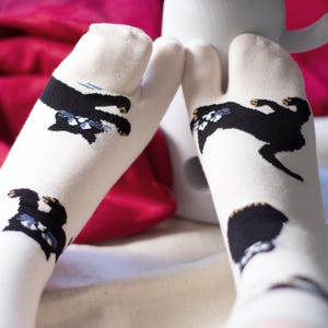 May include: White tabi socks featuring black cat designs with blue and tan details. These split-toe socks are intended for use with traditional Japanese footwear. A white mug and a pink surface are visible in the background.