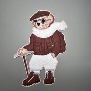 Può includere: Toppa ricamata raffigurante un orso in abbigliamento da alpinismo. L'orso indossa una giacca, un berretto e stivali marroni, con una sciarpa e pantaloncini bianchi. Porta un bastone da trekking e uno zaino, con un orologio al polso.