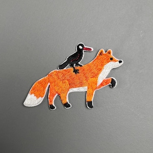 Può includere: Toppa ricamata raffigurante una volpe arancione con petto e coda bianchi, e un uccello nero con becco rosso appollaiato sulla schiena. La toppa ha un design dettagliato ed è probabilmente per abbigliamento o accessori.