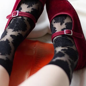 May include: A pair of deep red velvet Mary Jane shoes with silver-toned buckles, worn over sheer black socks featuring a dark, abstract, cloud-like pattern. The shoes have a rounded toe and a strap across the instep, showcasing a soft texture.