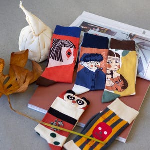 May include: A collection of colorful socks with unique designs. The socks feature illustrations of a woman's face, a man smoking a pipe, a person holding a squirrel, a panda, and a skull. The socks have various color schemes including red, blue, yellow, and black.