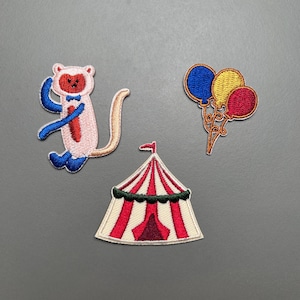 Puede incluir: Tres parches bordados: un mono rosa con detalles azules y rojos, una carpa de circo roja y blanca, y un grupo de tres globos azules, amarillos y rojos. Los parches están sobre un fondo gris.