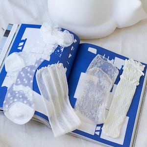 May include: An assortment of white socks with various designs, including polka dots, lace, and ribbed textures, are displayed on an open magazine with blue pages. A white decorative object is in the background. The socks are made of delicate materials.