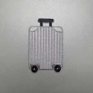 Puede incluir: Parche bordado de una maleta gris con ruedas y asa negras. La maleta tiene líneas verticales y está diseñada para parecerse a una pieza de equipaje. El parche está sobre un fondo gris.