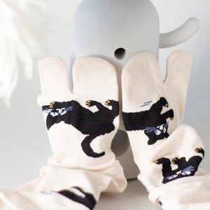 May include: Off-white tabi socks with a black cat design. The cat is depicted in a playful pose with yellow and blue accents. The socks are displayed in front of a gray, abstract sculpture.