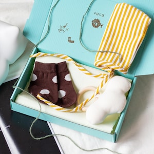 May include: A baby gift set in a light blue box, containing brown polka dot socks, a yellow and white striped headband, a white plush toy, and a wooden ring. The box is tied with a yellow and white striped ribbon and green twine. The box has the word "FUGU" printed on the lid.