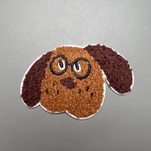 Peut inclure: Écusson en forme de tête de chien marron et beige avec des lunettes à monture noire et des oreilles marron foncé. L'écusson a un aspect texturé et flou et est souligné en blanc. Le nez et la bouche du chien sont également détaillés avec des coutures noires.