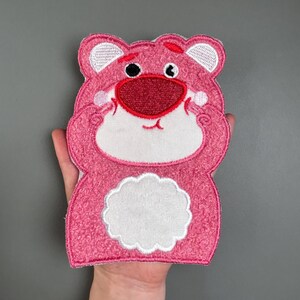 Könnte beinhalten: Rosa bestickter Aufnäher in Form eines Bären mit weißem Bauch und Gesicht. Der Bär hat eine rote Nase und Mund, schwarze Augen und weiße Akzente an den Ohren. Der Aufnäher ist eine Zeichentrickfigur.