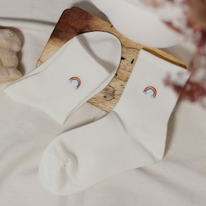 Könnte beinhalten: Zwei Paar weiße Socken mit Regenbogen- und Wolkenstickerei. Die Socken sind aus weichem Material und werden auf einer Holzoberfläche präsentiert. Das Regenbogen-Design hat rote, orange, gelbe, grüne, blaue und violette Farben.
