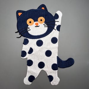 Può includere: Toppa ricamata a forma di gatto con testa e coda blu navy, occhi arancioni e baffi neri. Il corpo del gatto è bianco con pois blu navy. Il gatto sta salutando con una zampa.