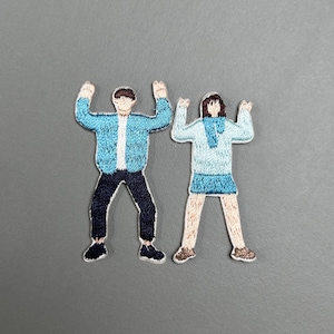 Könnte beinhalten: Zwei gestickte Aufnäher, die Figuren mit erhobenen Armen zeigen. Eine Figur trägt eine blaue Jacke, eine schwarze Hose und schwarze Schuhe. Die andere trägt einen hellblauen Pullover, einen blauen Rock und beige Schuhe. Die Aufnäher befinden sich auf grauem Hintergrund.