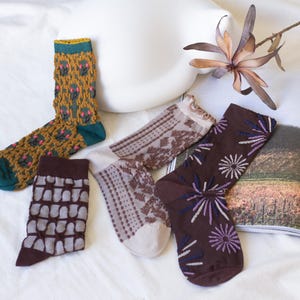Puede incluir: Un surtido de calcetines estampados en varios diseños y colores. Un calcetín presenta un patrón texturizado con detalles en verde, amarillo y rosa. Otros calcetines muestran patrones florales, geométricos y abstractos en tonos marrones, beige y morados.