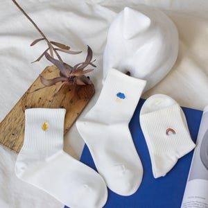 May include: Three pairs of white socks with embroidered designs. One sock features a yellow flame, another a blue cloud with rain, and the third a rainbow. The socks are ribbed and appear to be made of a soft material.