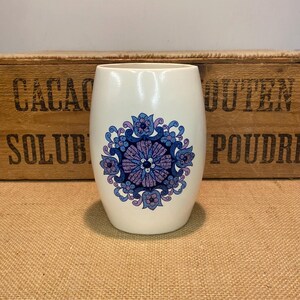 May include: A cream-coloured ceramic vase featuring a floral pattern in shades of blue and purple. The vase has a rounded shape and a smooth surface. The floral design is symmetrical and centered. A wooden crate is in the background.