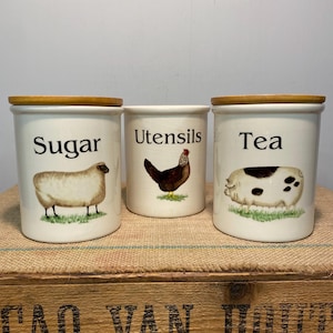 May include: Three ceramic kitchen canisters with wooden lids. The canisters are labelled "Sugar", "Utensils", and "Tea". Each canister features a different farm animal illustration: a sheep, a chicken, and a pig. The canisters are on a wooden surface.