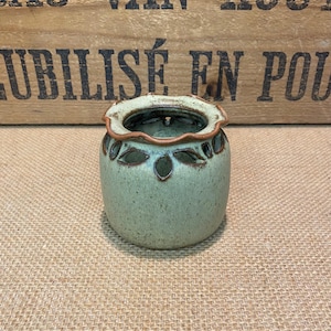 May include: A small, round, ceramic vase in a muted green hue. The vase features leaf-shaped cutouts and a scalloped rim with a brown accent. The interior is a darker green. A decorative piece for home decor.