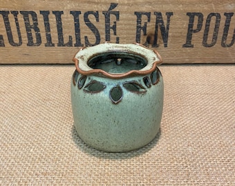 May include: A small, round, ceramic vase in a muted green hue. The vase features leaf-shaped cutouts and a scalloped rim with a brown accent. The interior is a darker green. A decorative piece for home decor.