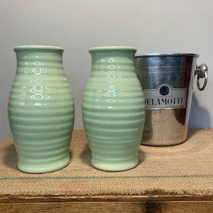 May include: Two pale green ceramic vases with horizontal ridges and a silver champagne bucket with the label "DELAMOTTE". The vases are cylindrical with a slightly flared top. The bucket has handles.