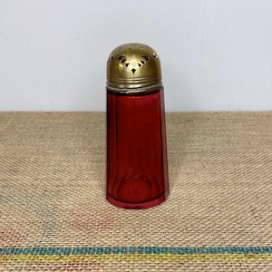 May include: A vintage red glass sugar dispenser with a brass lid featuring a decorative pierced design. The dispenser has a cylindrical shape with a slightly flared base.