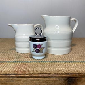 May include: A still life arrangement featuring a white ceramic egg cup decorated with blackberries, set between two white jugs. The jugs have a ribbed design and a handle. The egg cup has a silver lid.