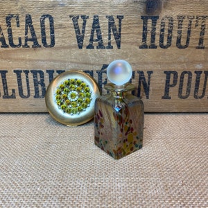 May include: A vintage gold compact with green rhinestone detailing and a decorative glass perfume bottle with a frosted glass orb stopper. The bottle is square and decorated with colourful spots.