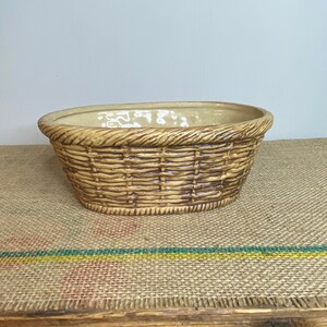 May include: A tan ceramic basket with a woven design. The oval-shaped basket has a textured rim and a light-coloured interior. The basket is sitting on a burlap surface.