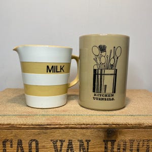 May include: Two vintage ceramic mugs with yellow and white stripes. One mug says "MILK" and the other says "KITCHEN UTENSILS." The mug with the utensils has a black and white illustration of kitchen utensils.