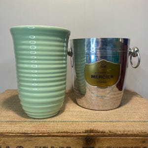 May include: A pale green ceramic vase with horizontal ridges stands beside a silver champagne bucket. The bucket has a gold label reading "Champagne Mercier Epernay". The bucket has two handles.